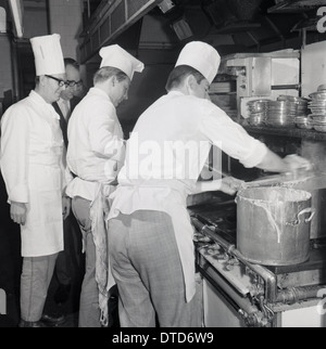Historical picture from 1960s showing professional chefs at a stove in a kitchen with hotel maitre d looking on. Stock Photo