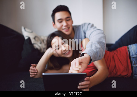 Relaxed teenage couple using touch screen computer while sitting on sofa at home. Young asian man and woman on tablet. Stock Photo