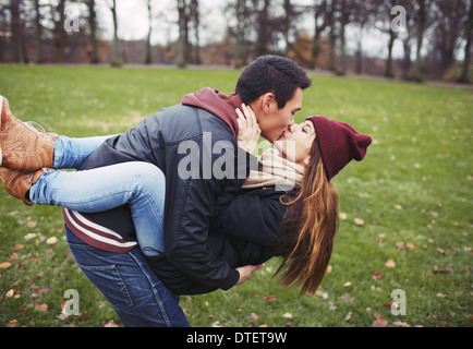 Attractive young man carrying his pretty girlfriend and kissing. Mixed race couple in love outdoors in park. Stock Photo