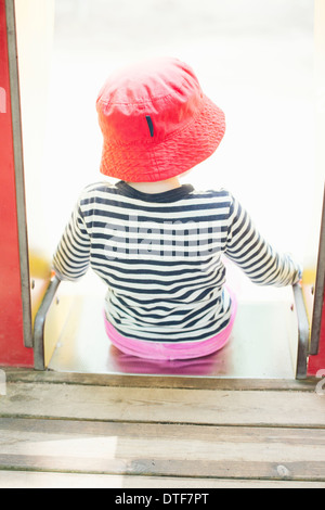 Childhood lifestyle moment. Little girl in read hat sitting at the top of a slide in a playground for children. Stock Photo