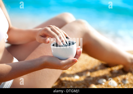 Young woman sitting on the beach and holding a cup of healing mud Stock Photo