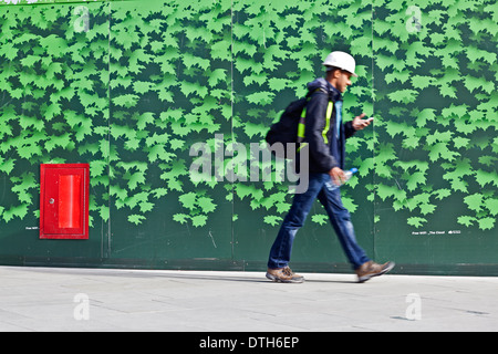 A construction worker using a mobile phone walks by architectural hoardings. Stock Photo