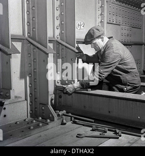 1950s, an historical picture showing a British workman wearing overalls and cap, with tools, checking the bolts on a steel beam. Stock Photo