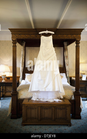 A full length white wedding dress hanging from a hanger on a four-poster bed England Stock Photo