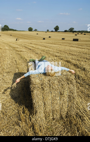 A woman lying on top of a square bale of packed straw at harvest time England Stock Photo