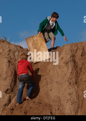 boys using old bit of wood to slide down sand dunes. Exmouth, Devon, UK Stock Photo