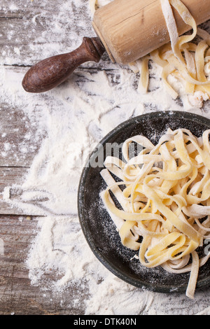 Top view on plane with raw homemade pasta with flour and vintage rolling pin over old wooden table. Stock Photo