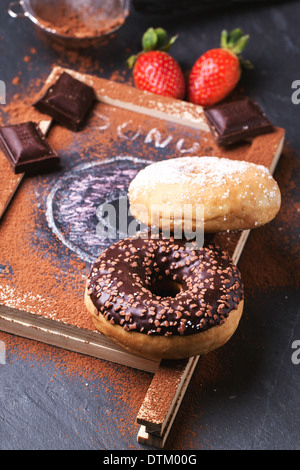 Sugar, chocolate and drawing donuts with fresh strawberries and dark chocolate served on chalkboard over black stone table. Stock Photo