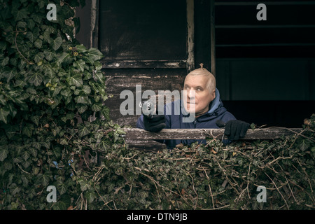 Masked senior escapee man aiming a gun, hiding on overgrown porch of old cabin. Stock Photo