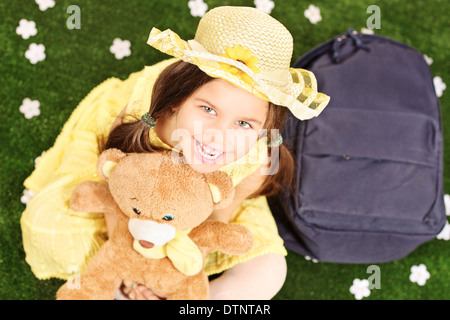 Cute little girl seated on green grass holding a teddy bear with backpack on the ground next to her Stock Photo