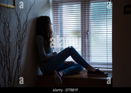 Young female with long hair wearing jeans, sad and looking towards a window, sitting in darkness at home. Side viewpoint. Stock Photo
