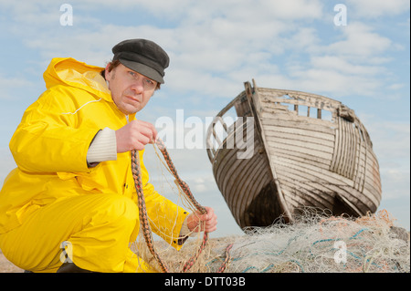 Fisherman in yellow waterproofs repairing fishing nets with an old wooden boat in the background. Stock Photo