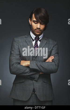 Young male business executive with his arms crossed looking down in thought. Thoughtful businessman in suit standing. Stock Photo