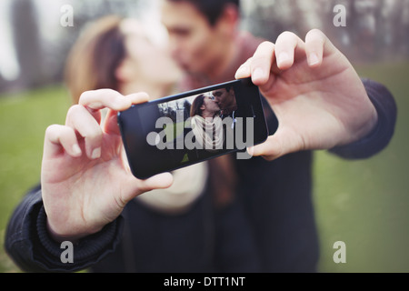 Loving young couple photographing themselves with a mobile phone while kissing at the park. Focus on smart phone. Stock Photo