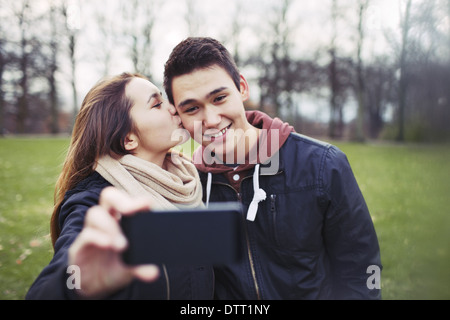 Pretty young girl kissing her boyfriend on cheeks while taking self portrait with a mobile phone. Mixed race couple in park. Stock Photo