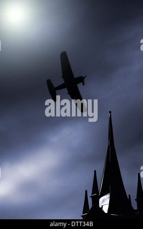 Old propeller fighter plane flying on a moonlit sky over building rooftop Stock Photo