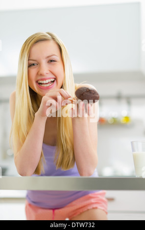 Happy young woman eating chocolate muffin in kitchen Stock Photo