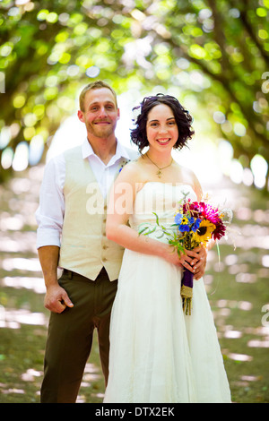 Bride and groom pose for a portrait in a backlit orchard on their wedding day in Oregon with light filtering through the trees. Stock Photo