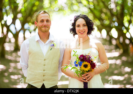 Bride and groom pose for a portrait in a backlit orchard on their wedding day in Oregon with light filtering through the trees. Stock Photo