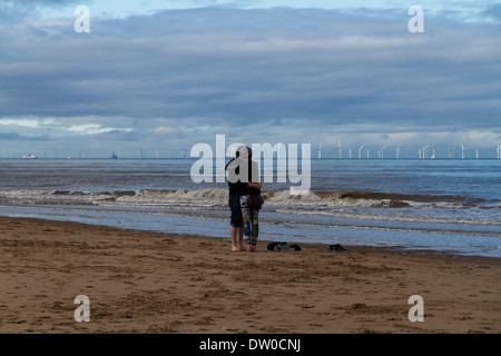 A couple hug on a beach with an offshore wind farm in the distance Stock Photo