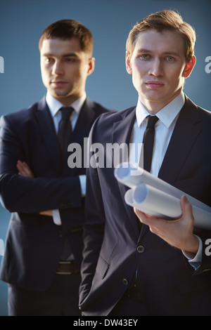 Vertical portrait of a young serious architect with drafts looking at camera while his colleague standing on the foreground Stock Photo