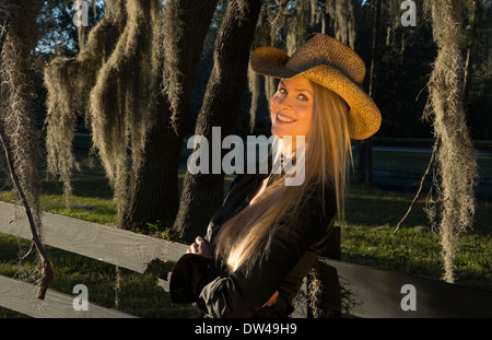 attractive 50 year old blonde woman cowgirl with a hat outdoors leaning against fence with spanish moss Stock Photo