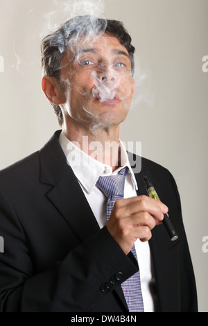 Middle aged confident man enjoying smoking an e-cigarette or vaporizer exhaling a cloud of smoke and peering through the fumes Stock Photo