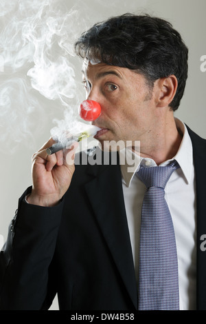 Middle-aged businessman wearing a red nose like a clown puffing on an e-cigarette standing sideways peering through the fumes. Stock Photo