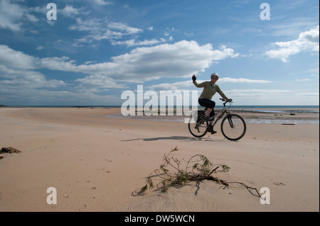 Woman cycling and waving on the beach of Beadnell Bay, Northumberland, England, on a sunny day Stock Photo