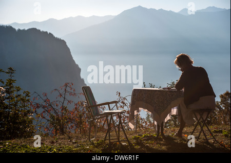 A woman sitting at a table, high in the mountains, writing a letter in the early morning light. Stock Photo