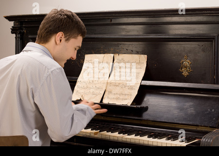 View from the rear of a young man smiling as he plays the piano using an old vintage music score on an upright wooden piano Stock Photo