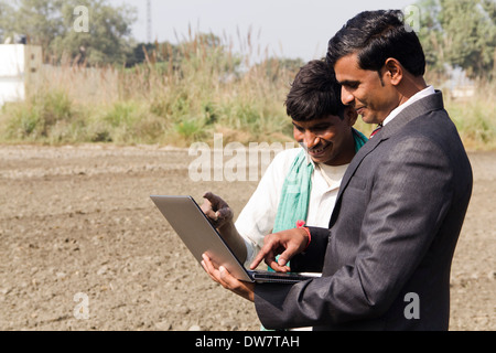 farmer working in plowedfield and busineeman standing with laptop Stock Photo