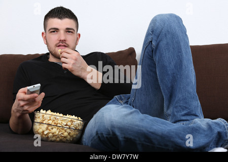 Young man lying on a sofa, watching tv and eating popcorn Stock Photo