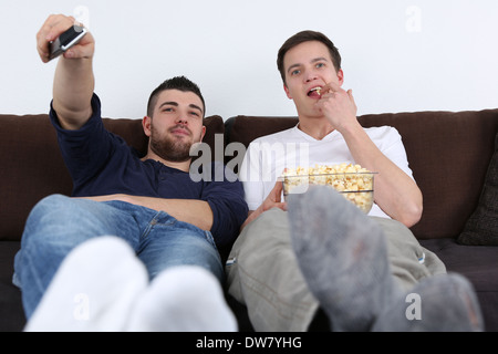 Young people sitting on a sofa, relaxing and eating popcorn while watching tv Stock Photo