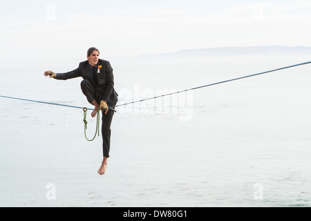 A groom in a suit goes to stand on a slackline above water to walk to his bride to be. Stock Photo