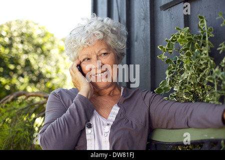 Happy elderly woman sitting on a bench in backyard talking on mobile phone and smiling Stock Photo