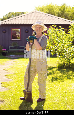Full length portrait of active senior woman wearing hat with gardening tools smiling outdoors. Elder woman standing with shovel. Stock Photo