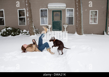 Mid adult woman lying in snow outside house playing with dog Stock Photo