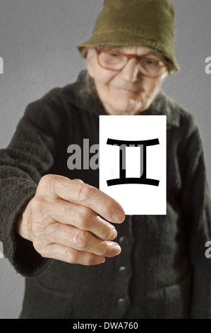 Elderly woman holding card with printed horoscope Gemini sign. Selective focus on card and fingers. Stock Photo