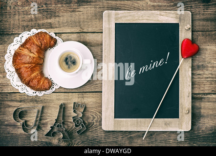 Cup of black coffee with croissant, blackboard and heart decoration on rustic wooden background. Stock Photo