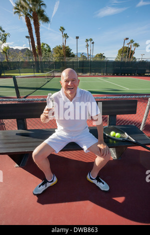 Portrait of happy senior male tennis player with water bottle relaxing on court Stock Photo