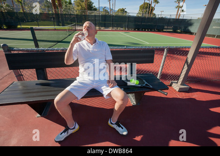 Senior male tennis player drinking water while relaxing on court Stock Photo