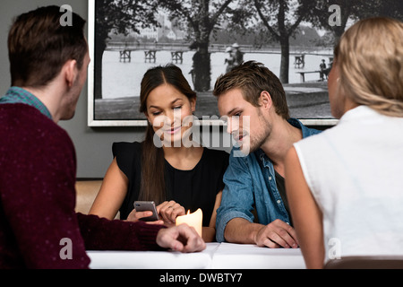 Four young adult friends sitting in cafe looking at cellphone Stock Photo