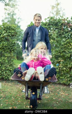 Playful father pushing daughters on wheelbarrow at yard Stock Photo