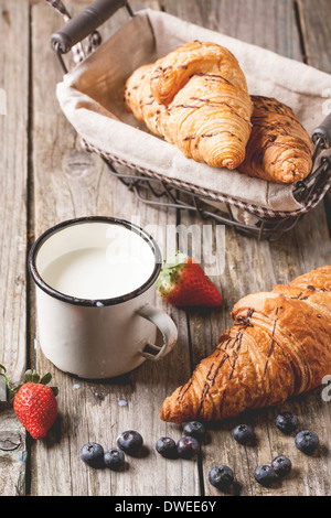 Fresh croissant with vintage mug of milk and berries over old wooden table. Stock Photo