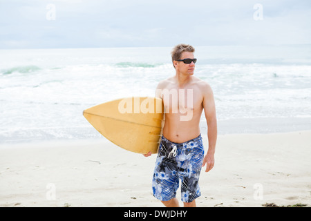 Serious looking male surfer walking on beach while carrying surfboard Stock Photo