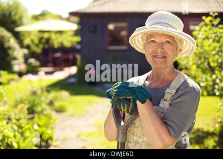 Portrait of senior woman wearing hat with gardening tools outdoors. Elder woman standing with shovel in her backyard garden Stock Photo