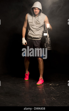 Young boxer arriving for a training session carrying has gloves by the laces in his bandaged hands as he approaches out of the darkness. Stock Photo