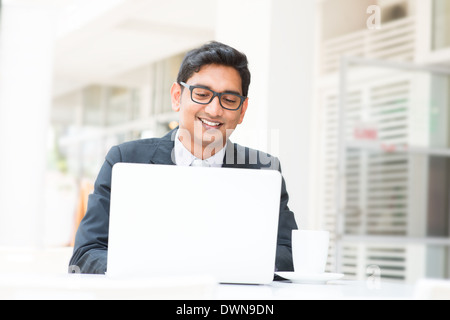 Young Asian Indian businessman using a notebook computer or laptop during office break at cafe, relaxing with a cup of coffee. India male business man, real modern office building as background. Stock Photo