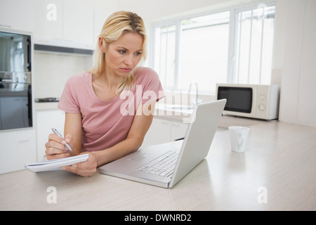 Woman using laptop while writing notes in kitchen Stock Photo